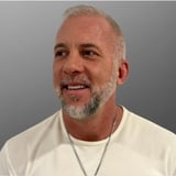 Middle-aged man with gray hair and facial hair wearing a white t-shirt and chain necklace against a gray background