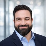 Professional headshot of a man with dark hair and full beard wearing a navy blazer and light blue shirt against blurred office background