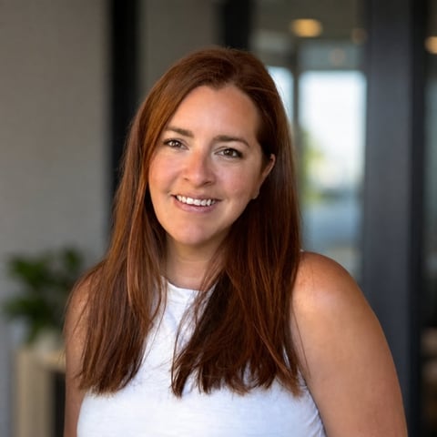 Woman with long reddish-brown hair wearing a white sleeveless top, smiling at camera in an indoor office setting
