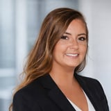 Professional headshot of a woman with long brown hair wearing a black blazer over a white top, smiling at the camera against a blurred office background